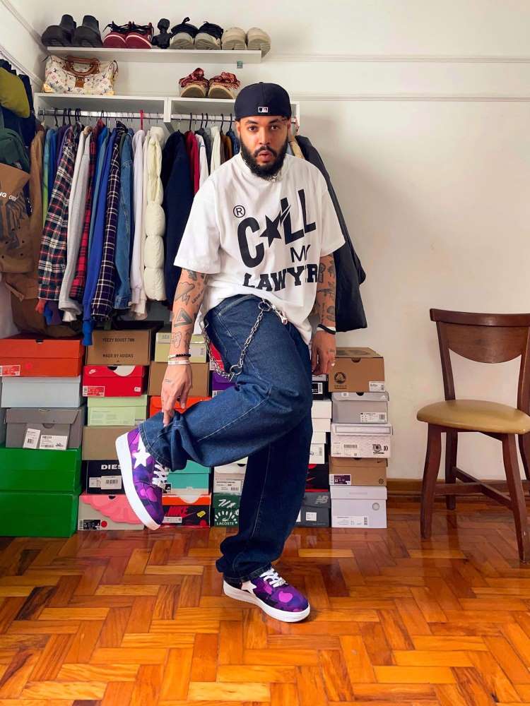 Homem de pele negra usando blusa oversized, boné pra trás, calça jeans com corrente e tênis roxa