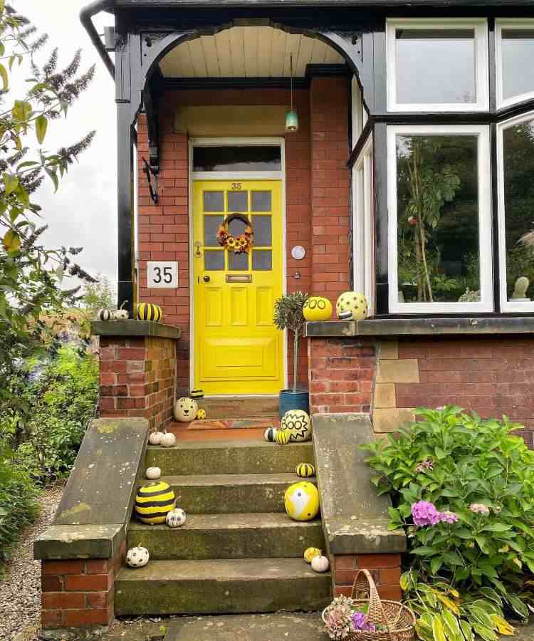 Entrada de casa com parede de tijolos à vista, porta amarela com guirlanda de girassol e abóboras amarelas com rosto, frases e desenhos 