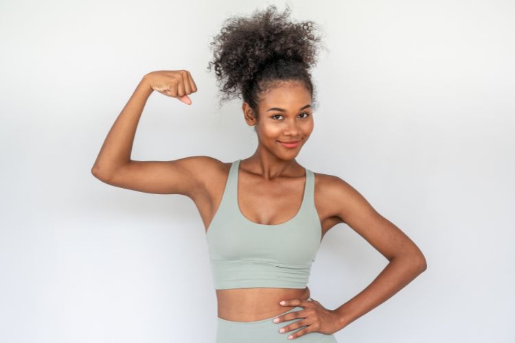 A imagem mostra uma pessoa posando em frente a um fundo branco, exibindo seu bíceps. Ela está vestindo um top esportivo verde claro e fazendo uma pose de força, com um braço flexionado para mostrar o bíceps. O fundo é completamente branco, colocando foco total na pessoa em primeiro plano.