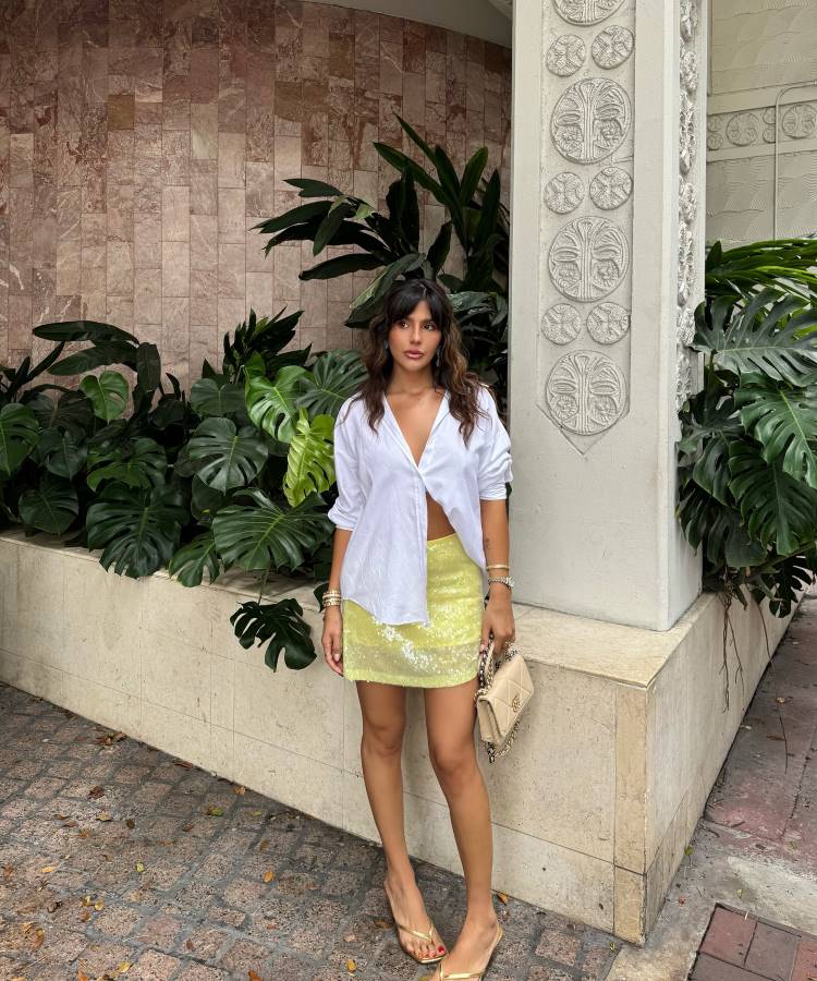 Uma mulher posa de pé em frente a folhagem exuberante. Ela veste uma camisa branca amarrada, uma minissaia de paetês amarelos e chinelos dourados. Ela tem uma bolsa creme pendurada no ombro e pulseiras.








