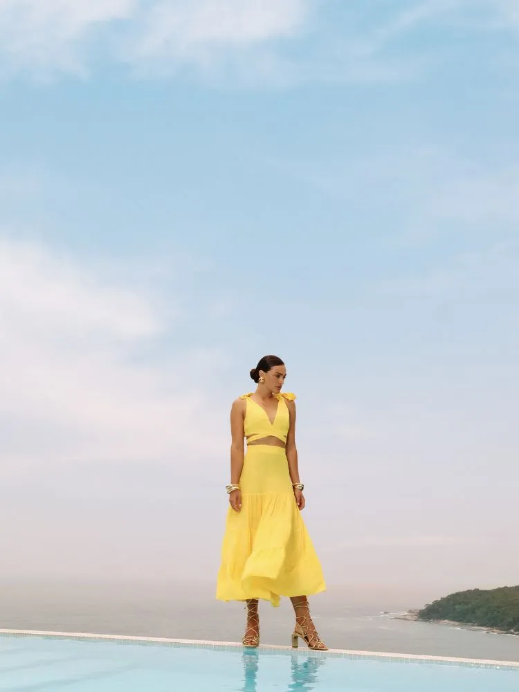 Uma modelo posa em uma rocha com o mar ao fundo, o céu está em um degradê de rosas e azuis. Ela Usa um conjunto de saia e top amarelo fluído da coleção Dias Dourados da DIMY