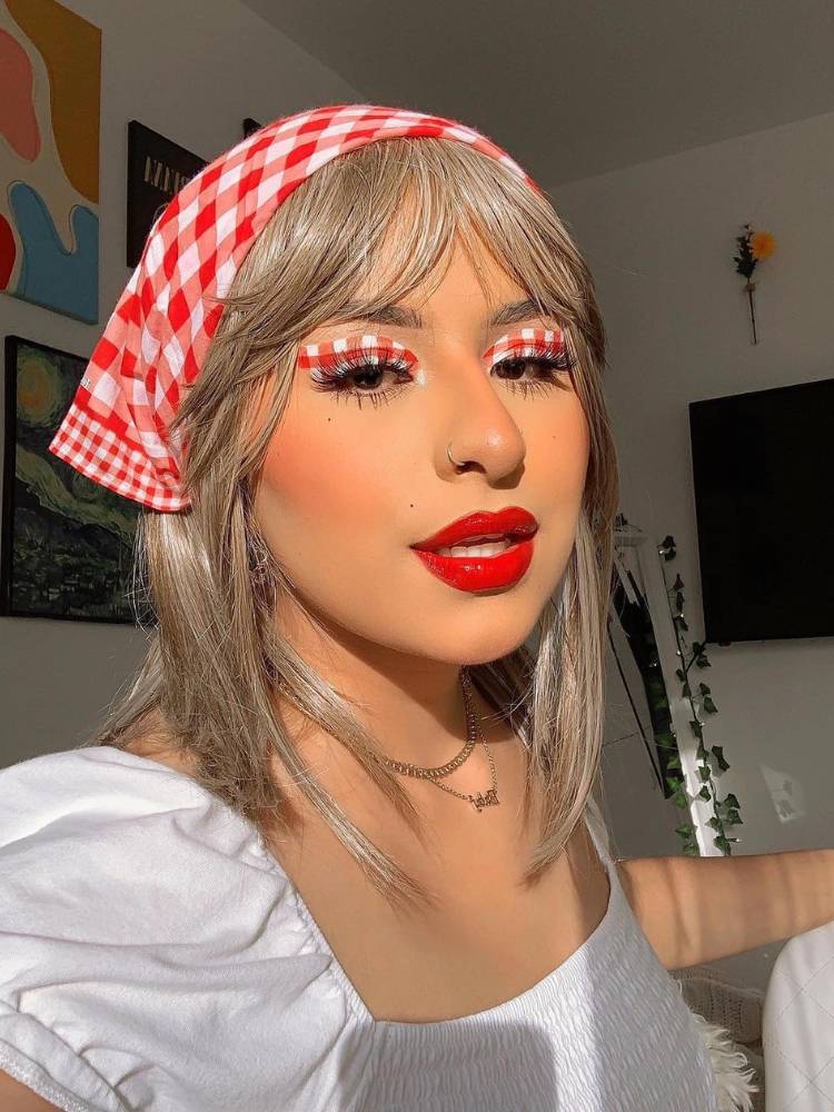 Mulher de pele clara usando blusa branca, maquiagem xadrez vermelha e branca e bandana xadrez vermelha e branca no cabelo solto
