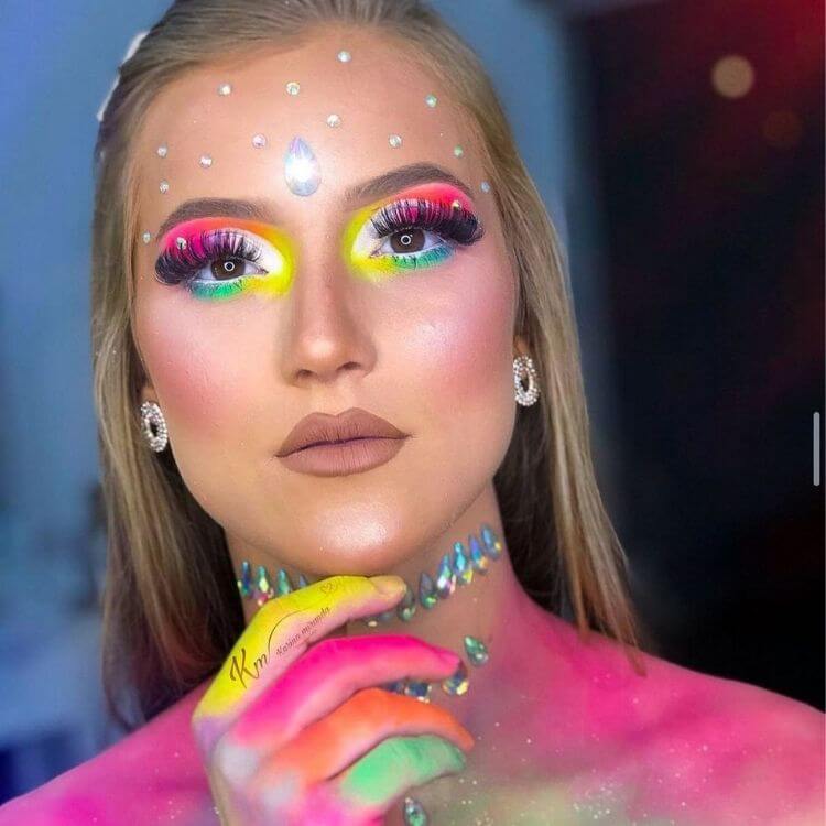 Mulher de pele clara usando maquiagem neon colorida nos olhos e corpo, pedrarias na testa e no pescoço e batom nude