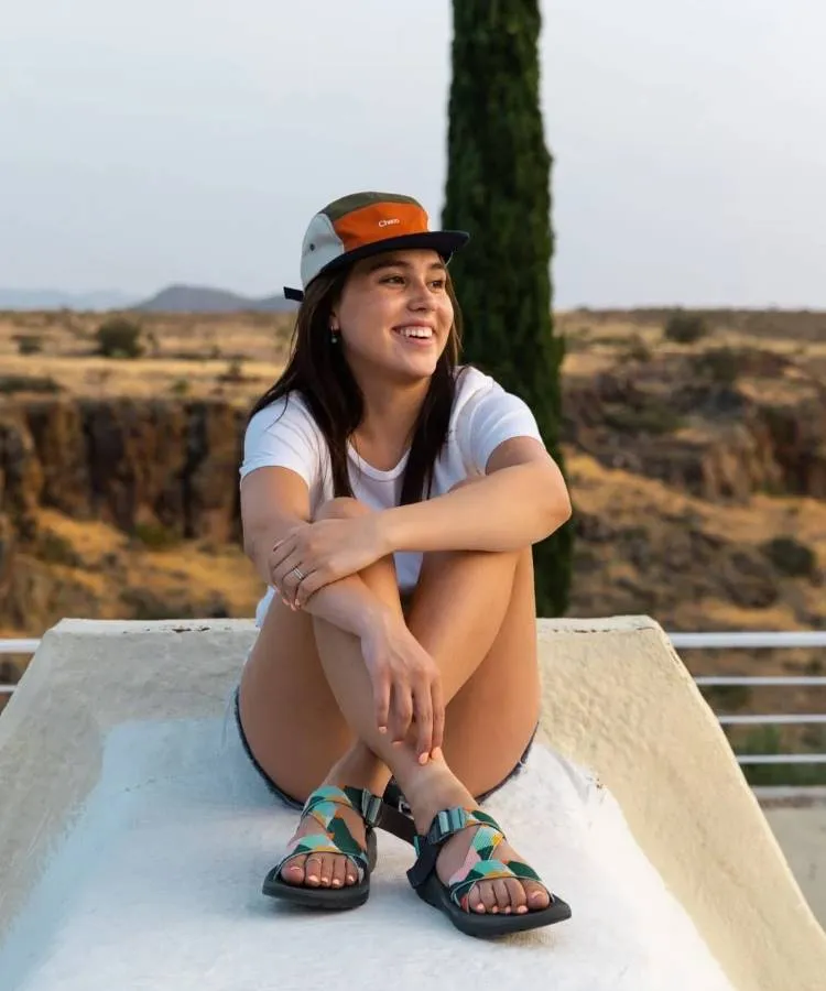 A foto mostra uma mulher branca de cabelos pretos sentada em um banco de concreto, ela usa um boné, camiseta branca e shorts jeans. Nos pés uma papete de estampas azuis
