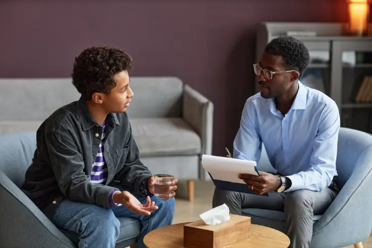 Dois homens de pele negra sentados à mesa com caixa de papel, sendo um mais velho com prancheta nas mãos e o outro segurando um copo de água