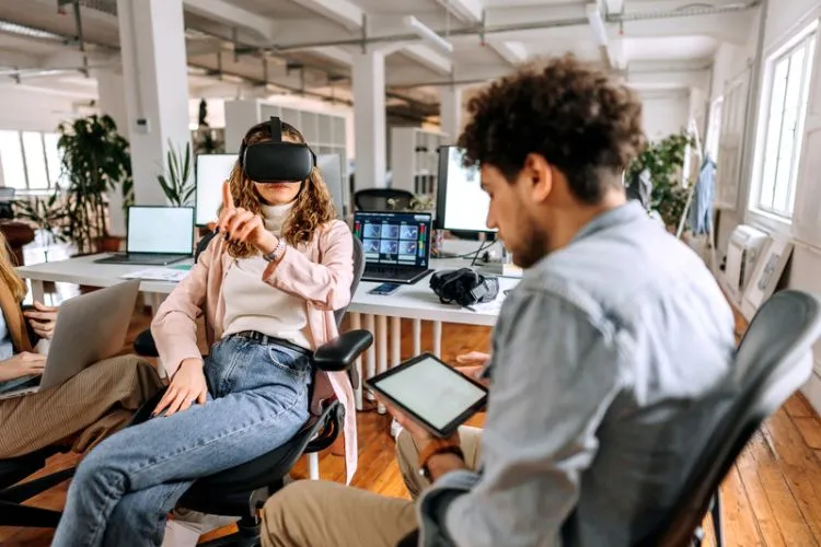Foto de mulher usando óculos de realidade virtual ao lado de designer de games segurando tablet e fazendo teste