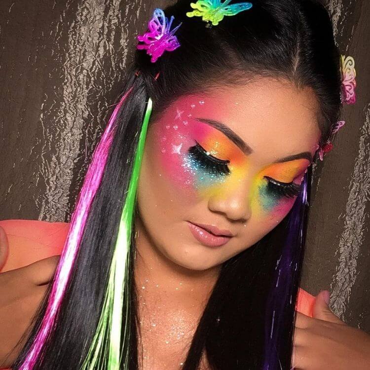 Mulher de pele clara com maquiagem colorida, adesivos prateados, glitter e mechas colorida no cabelo 