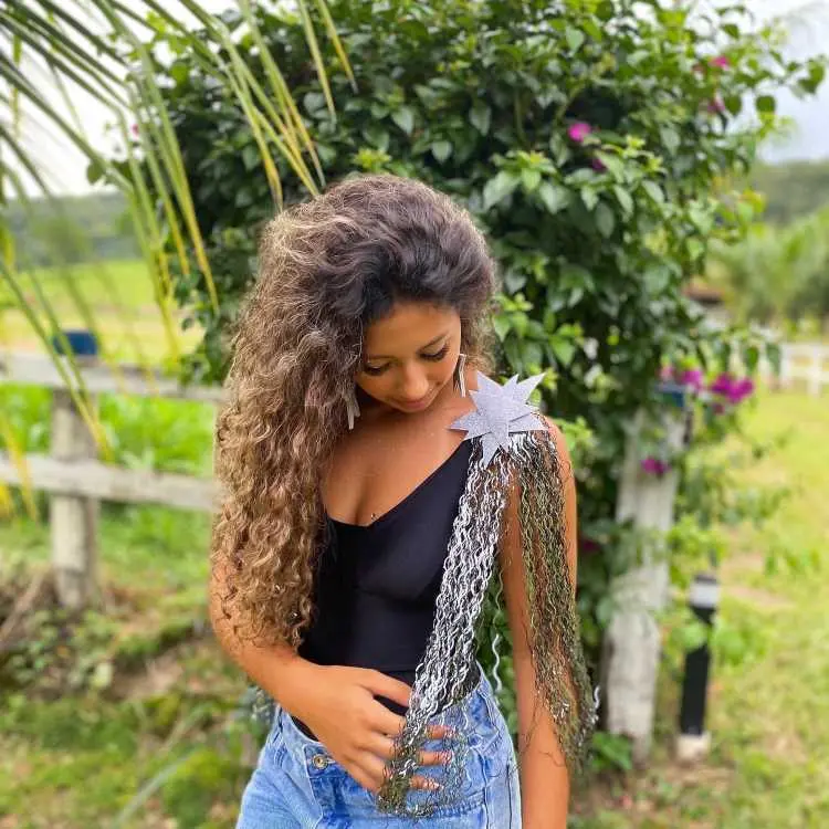 Uma mulher com cabelos cacheados longos veste um body preto e shorts jeans. Ela usa uma ombreira prateada em forma de estrela com franjas metálicas. Ao fundo, há plantas, flores e uma paisagem campestre iluminada.