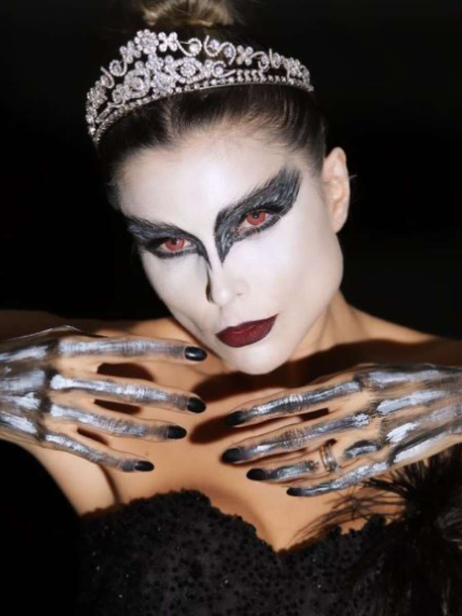Imagem em fundo preto com Flávina Viana no centro. A foto foca no rosto dela, que possui maquiagem de asas pretas nos olhos. Ela usa ainda coroa na cabeça e suas mãos também estão pintadas.