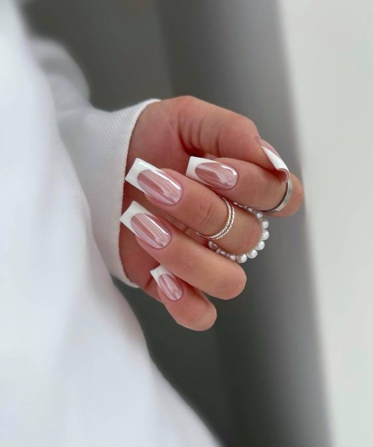 Unhas ovais com esmalte nude cintilante em mãos delicadas adornadas com anéis minimalistas. Fundo suavemente desfocado destaca a beleza das mãos e das unhas, evocando elegância discreta. Uma pulseira fina complementa o visual sofisticado, perfeito para noivas modernas.



