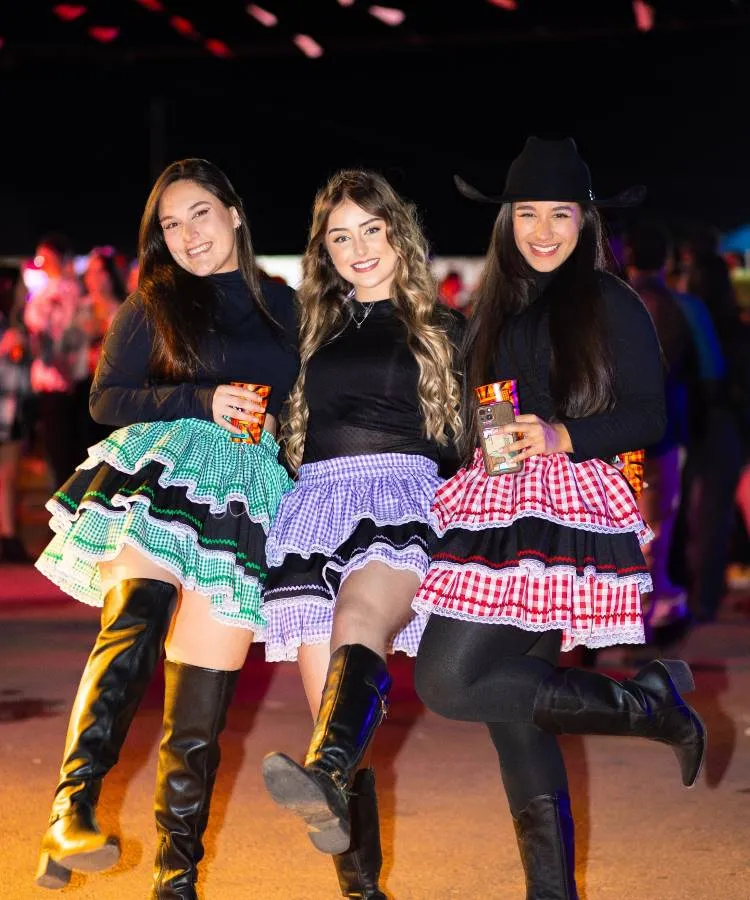 Três mulheres em trajes tradicionais festivos, sorrindo e levantando uma perna. Usam blusas pretas, saias coloridas de estampas quadriculadas e botas. Estão em um evento noturno ao ar livre, segurando bebidas, com luzes coloridas e ambiente animado ao fundo.
