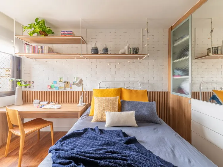 Vista para parede da cabeceira da cama do quarto da irmã mais velha. Prateleira acima com livros, objetos e planta. Ao lado da cama, uma mesa com cadeira para estudos.