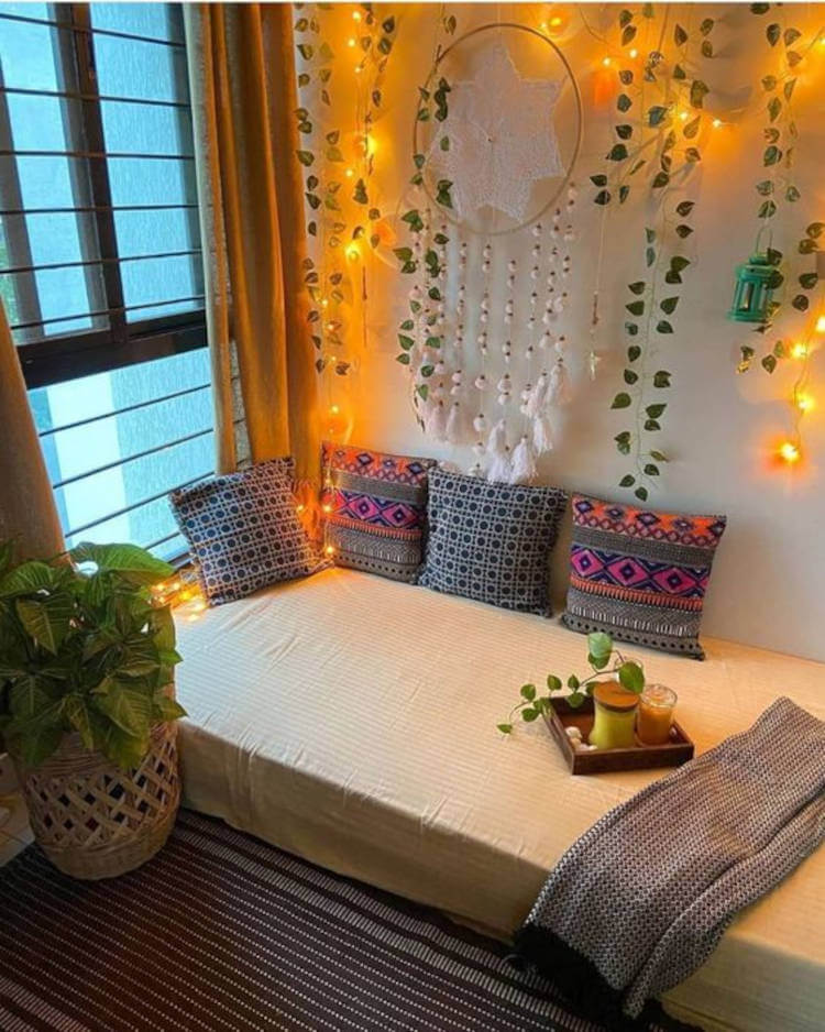 Cantinho zen com colchão no chão, almofadas coloridas, vaso de plantas, plantas pendentes, coberta e iluminação amarela.