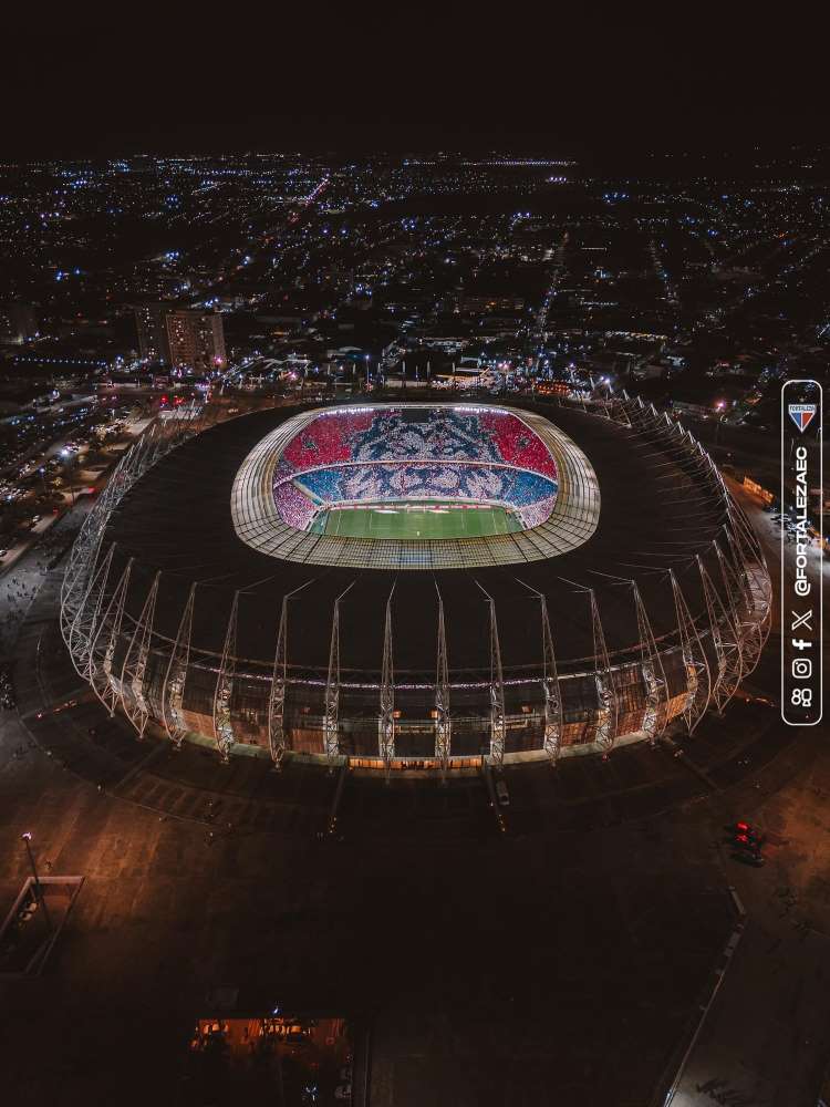 Foto tirada à noite da Arena Castelão iluminada e com mosaico de Leão na arquibrancada.