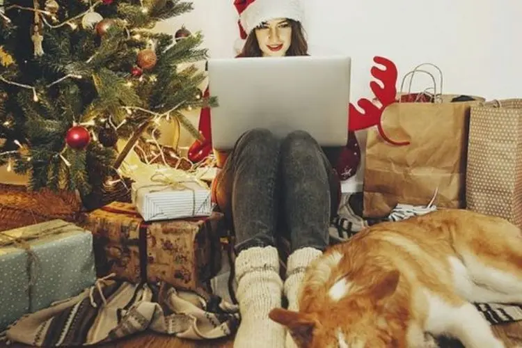 Mulher de pele clara usando calça jeans, sueter vermelho, gorro do papai Noel, sentada ao lado de árvore-de-Natal, presentes e cachorro, segurando um notebook