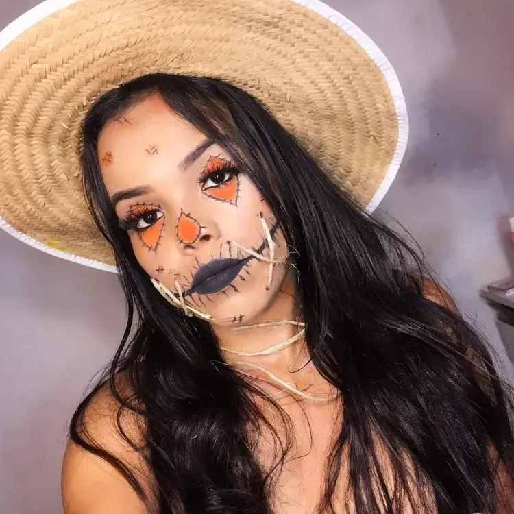 Foto de mulher de pele clara com chapéu de palha, maquiagem de espantalho com efeito de remendo nos olhos e nariz, além de boca com impressão de costurada com delineado e corda
