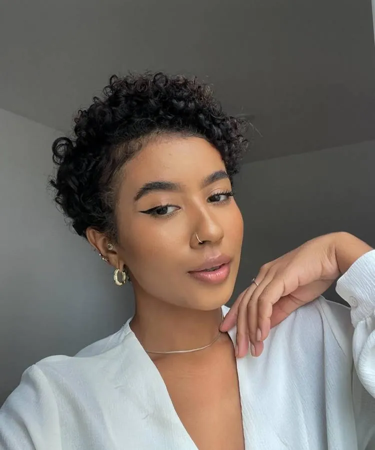 Uma mulher com cabelo curto e encaracolado posa, olhando para a câmera com maquiagem nos olhos e batom. Ela usa brincos, um anel no nariz e um colar simples, vestindo uma blusa branca.