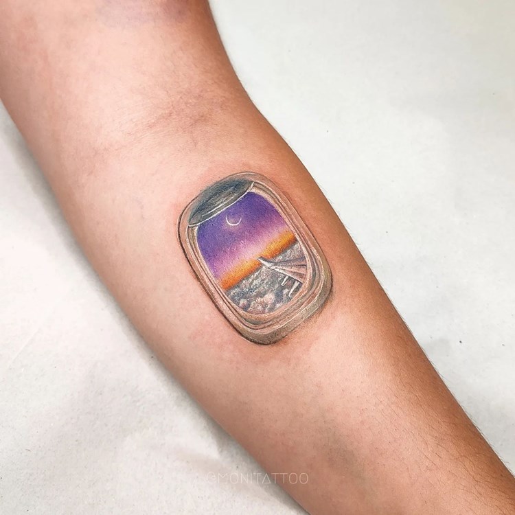 Tatuagem de viagem no braço de janela de avião com vista para a asa e um céu sobre as nuvens