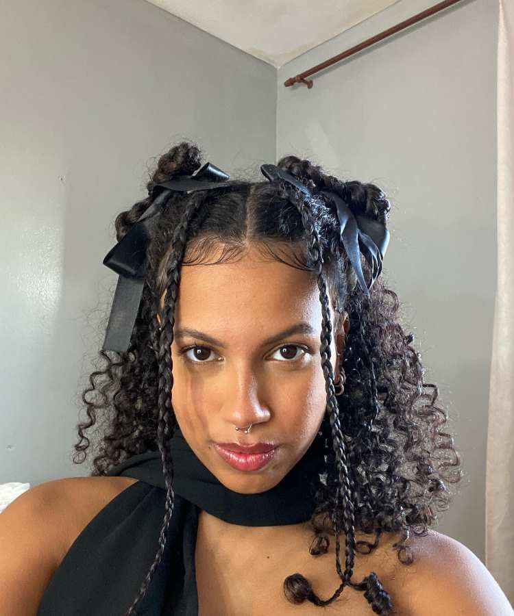 Penteado com cabelo cacheado e tranças finas, decorado com dois laços pretos grandes, em ambiente interno bem iluminado.