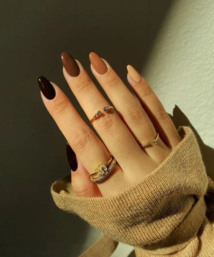 A imagem mostra uma mão com unhas longas e pontiagudas, pintadas em tons de marrom, variando do claro ao escuro. Uma unha marrom, mais escura, destaca-se entre as outras. A mão está adornada com anéis delicados e parcialmente coberta por uma manga de malha bege.

