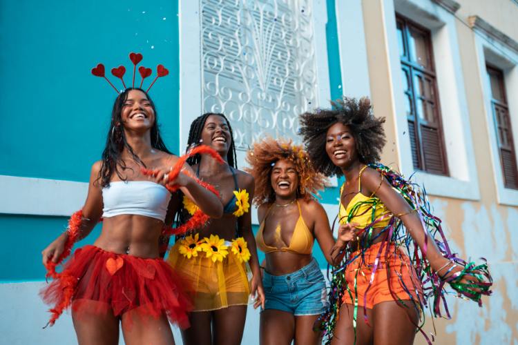 Quatro mulheres alegres celebrando o Carnaval em frente a construções coloridas. Usam roupas vibrantes, acessórios temáticos e sorrisos contagiantes, transmitindo energia, diversidade e espírito festivo. O cenário reflete a cultura e a alegria das festas de rua brasileiras.