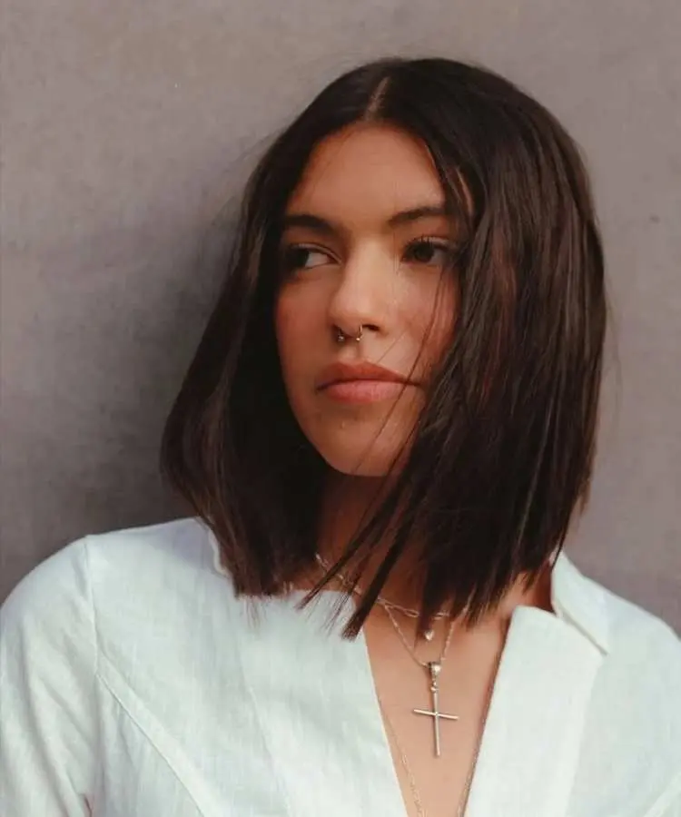 Mulher com cabelo médio castanho escuro, liso e corte reto na altura dos ombros, repartido ao meio. Ela usa blusa branca, colares delicados com pingente de cruz e piercing septo. Fundo cinza neutro destaca o visual moderno.