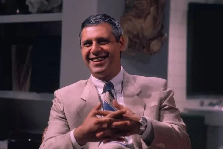Homem de cabelos curtos grisalhos, sorridente, veste terno bege, camisa branca e gravata azul com faixas diagonais. Está sentado em ambiente interno com decoração neutra ao fundo. Membro do elenco de "Vale Tudo", hoje icônico por seu papel inesquecível.