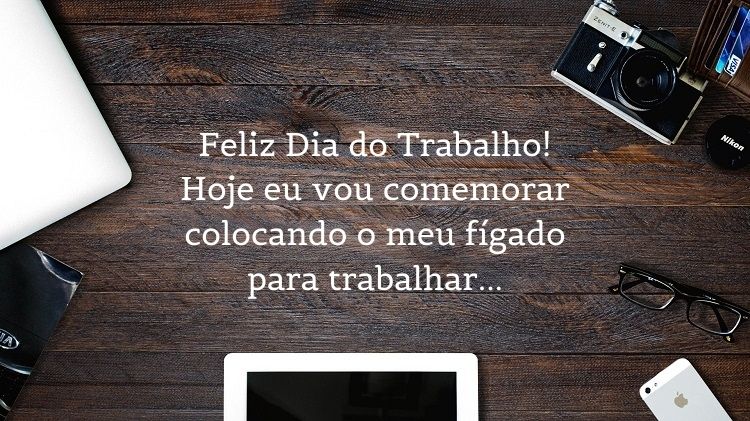 Foto de mesa de madeira com tablet, celular, computador, óculos, máquina fotográfica e frasr "Feliz Dia do Trabalho! Hoje eu vou comemorar colocando o meu fígado para trabalhar…"