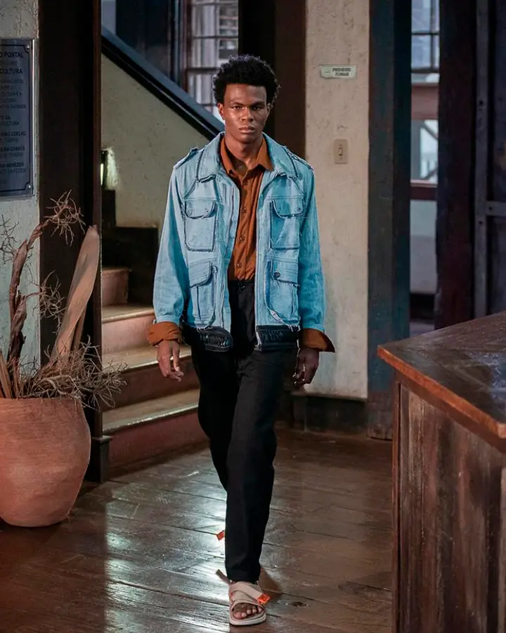 Modelo masculino usa blusa marrom, calça preta e jaqueta azul.