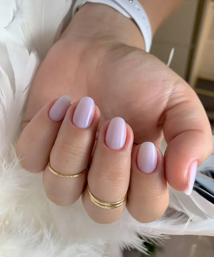 Unhas elegantes, claras, curtas, no formato amendoada. A mão da mulher possui uma corrente, possivelmente de um relógio, e ela usa anéis dourados. Sua mão está próxima a penas brancas.
