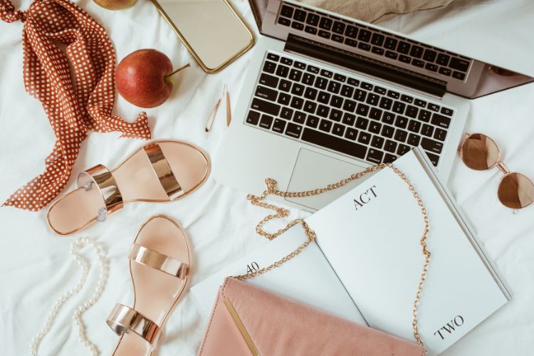 Mesa com laptop, acessórios elegantes, sandálias douradas, óculos de sol, livro e itens de moda sobre tecido branco. A composição ilustra lifestyle e criatividade, destacando o contexto de discussão sobre a diferença entre influencer e blogueira no mundo digital.





