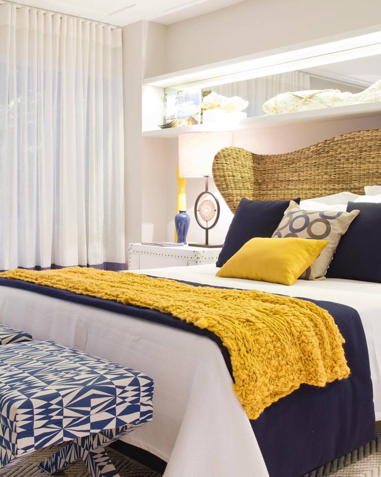 Decoração de verão de um quarto elegantemente decorado. Há uma cama grande com lençóis brancos, cobertor azul escuro e uma manta amarela mostarda. Vários travesseiros estão na cama, incluindo dois grandes travesseiros azuis escuros e um travesseiro amarelo mostarda. Uma mesinha de cabeceira branca está ao lado da cama com um abajur moderno. As cortinas brancas semi-transparentes cobrem a janela ao lado da cama, permitindo que a luz natural entre no quarto. Acima da cabeceira da cama há prateleiras embutidas iluminadas contendo itens decorativos como vasos e caixas. O quarto é iluminado e apresenta uma paleta de cores suaves.


