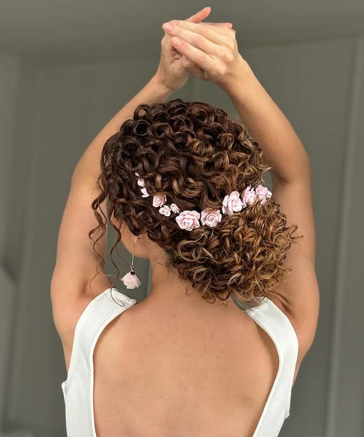 Penteado com coque baixo de cachos castanhos, decorado com flores rosadas, visual de noiva em vestido branco aberto nas costas.