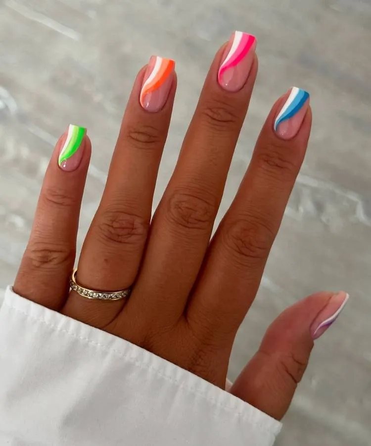 A imagem mostra um close-up de uma mão com os dedos estendidos, exibindo unhas pintadas com um design multicolorido e brilhante. Cada unha tem uma combinação diferente de cores em um padrão que lembra mármore ou aquarela. O polegar tem verde e branco, o indicador laranja e branco, o dedo médio rosa e branco, o anelar azul e branco, e o dedo mínimo roxo e branco. A mão está adornada com um anel dourado no dedo anelar. O fundo é desfocado, mas parece ser uma superfície cinza que pode ser interpretada como mármore ou concreto.
