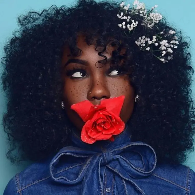 Frase para foto pensativa mulher negra com cabelo cacheado usa flores no cabelo, usa rosa na boca e veste camisa jeans com laço no pescoço Frase para foto pensativa mulher negra
