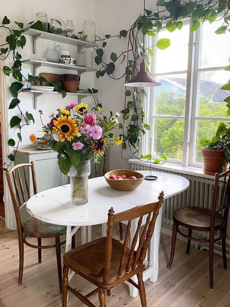 Copa com mesa branca, cadeira de madeira, janela com vista para montanha e diversas plantas na decoração, pendentes e em vasos