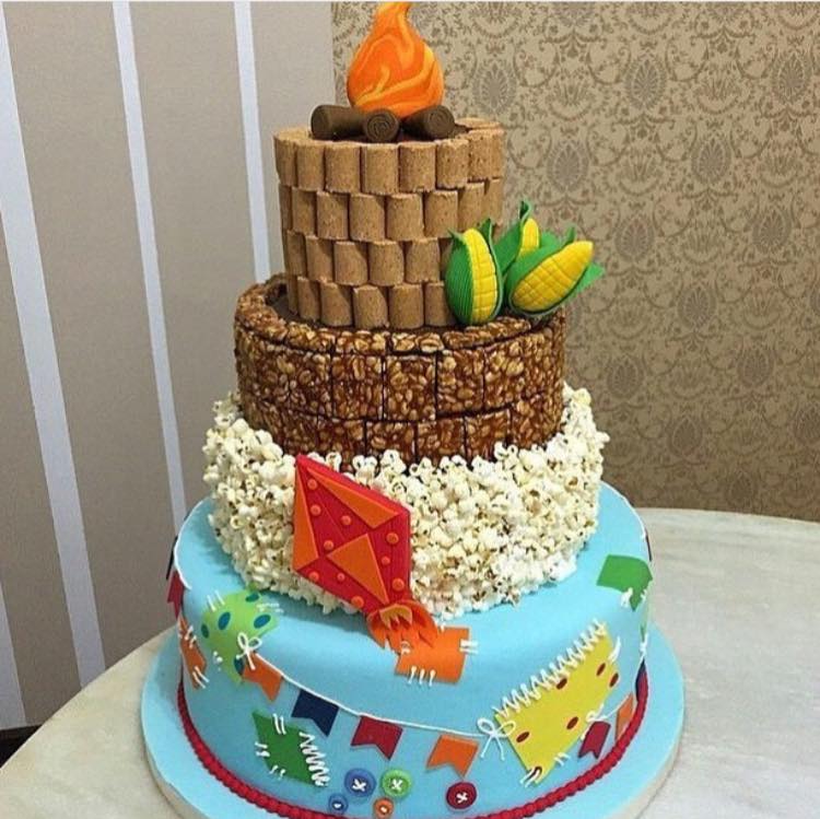 A imagem representa decorações de festa junina: bolo de são joão com pipoca, pé de boleque e paçoca