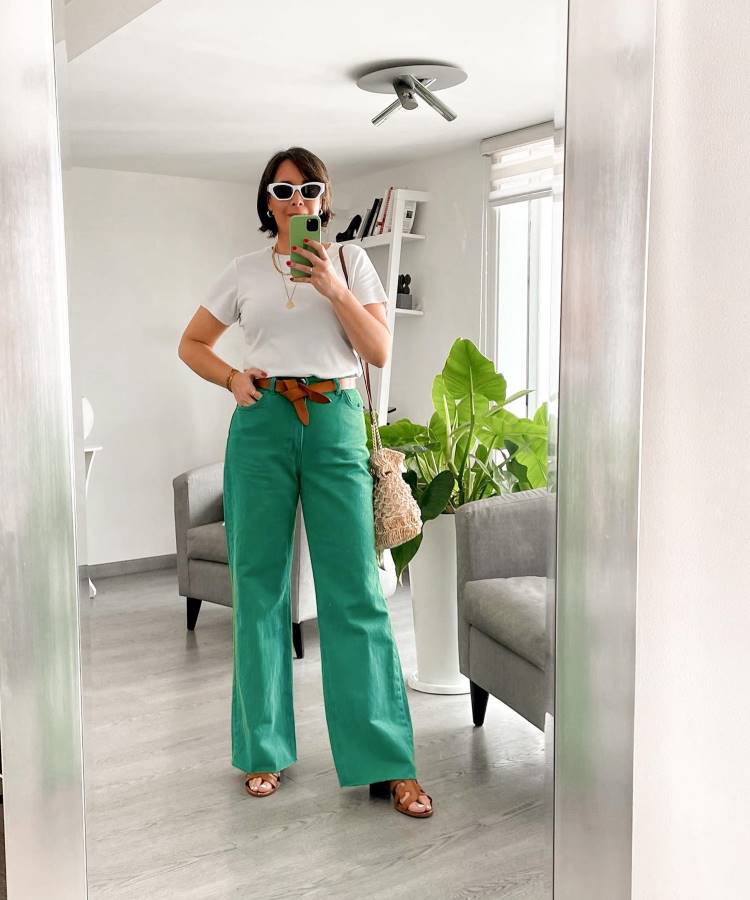A imagem mostra uma mulher vestindo uma calça verde wide leg com cinto marrom, combinada com uma camiseta branca básica. Ela complementa o look com sandálias marrons, óculos de sol brancos e uma bolsa de crochê, criando um estilo casual moderno.