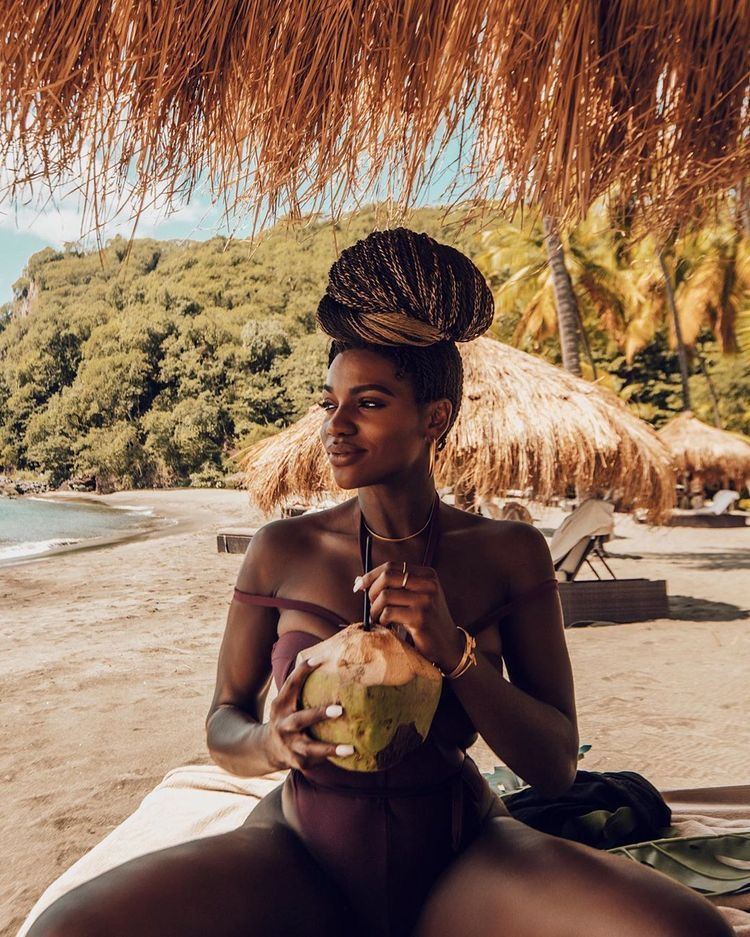 Mulher de pele negra usando maiô marrom, sentada tomando água de coco na areia à beira do mar 