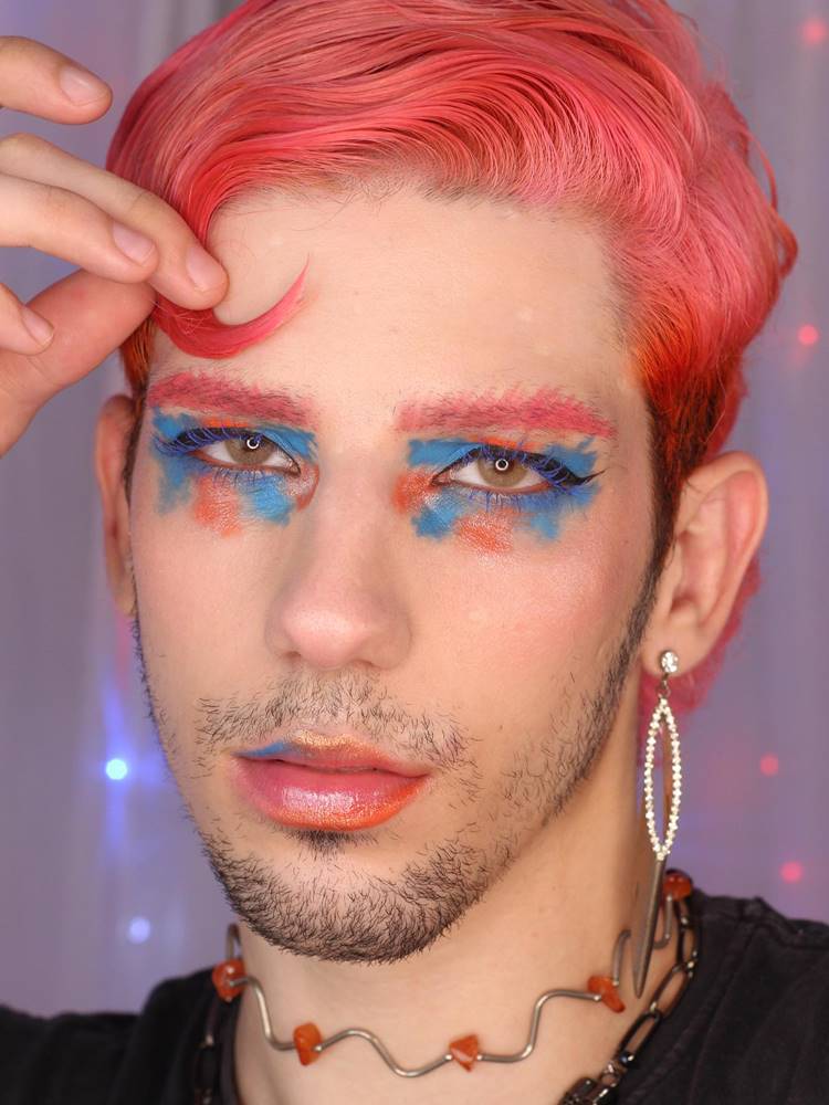 Homem de cabelo rosa com make azul e laranja nos olhos + delineado preto. Nos lábios, ele usa as mesmas sombras para contornar