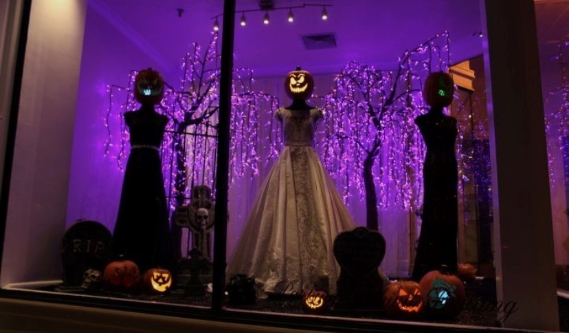 Vitrine de loja de roupa de noiva com decoração de Halloween, com manequins com cabeça de abóbora iluminada por dentro 