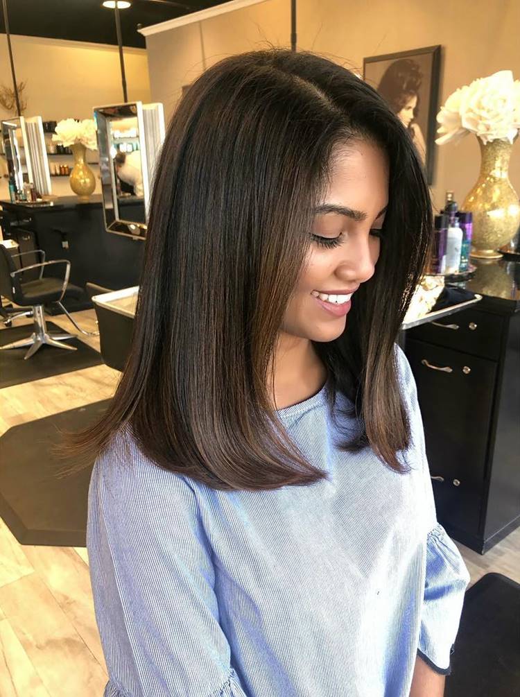 Mulher sorridente com cabelo liso, cortado na altura dos ombros, usando blusa azul clara com mangas largas. Ela está em um salão de beleza, com espelhos e produtos ao fundo, criando um ambiente elegante e acolhedor. A iluminação é suave.