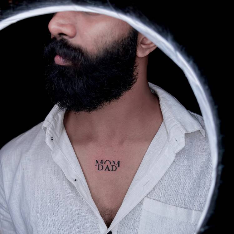 A imagem apresenta um close-up de um homem barbudo vestindo uma camisa branca aberta, revelando uma tatuagem preta no centro do peito, que diz "MOM" em cima de "DAD". A iluminação forma um círculo ao redor da cabeça do homem, com o fundo escuro.







