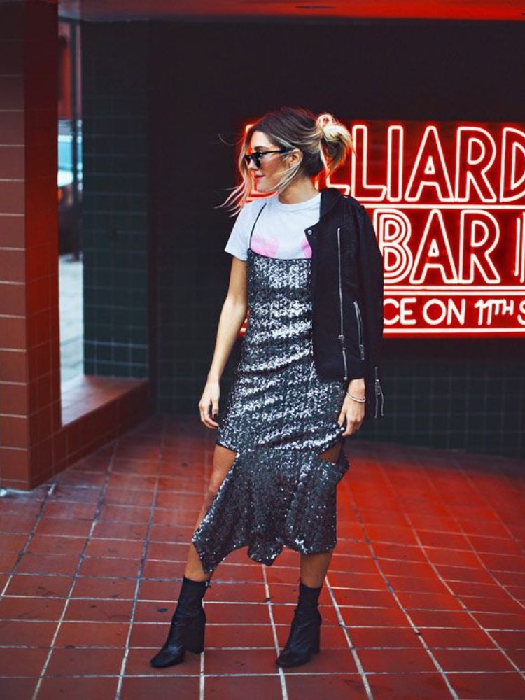 Mulher de pele clara com bota preta, vestido metalizado e blusa por baixo