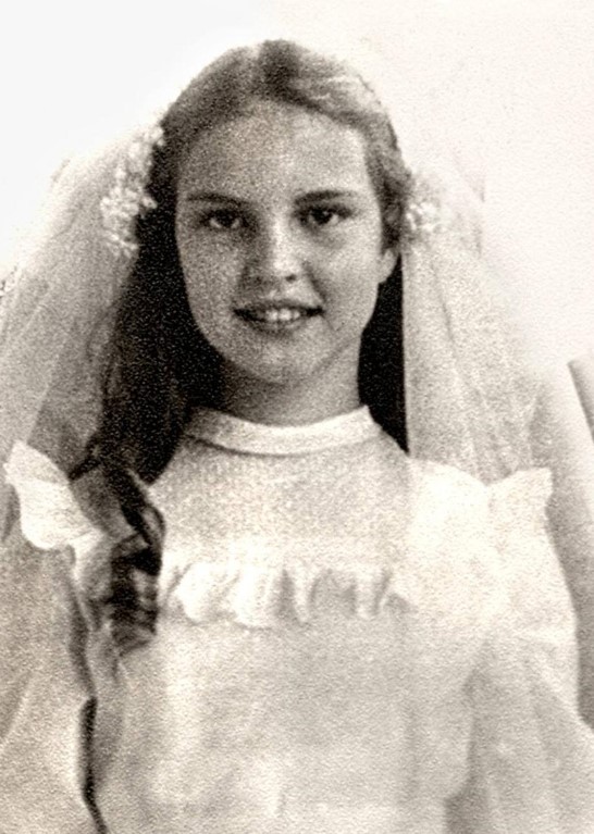 Foto em preto e branco de Elke Maravilha criança com um vestido branco com babados e um véu branco preso ao cabelo. Retrato em preto e branco de Elke Maravilha quando criança, sorrindo, com um vestido branco com babados e um véu branco preso ao cabelo.