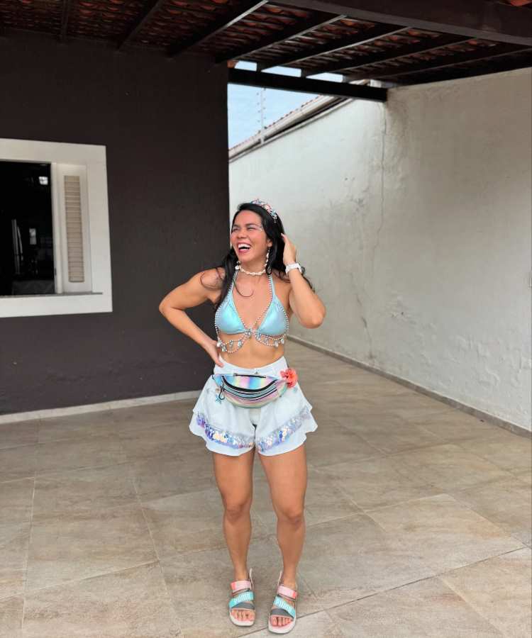 Mulher jovem sorridente com cabelo longo e tiara posa em varanda coberta. Veste top azul brilhante, short branco com detalhes holográficos, pochete colorida e sandálias azul-claras. Fundo com paredes brancas e janela destaca ambiente casual e ensolarado.