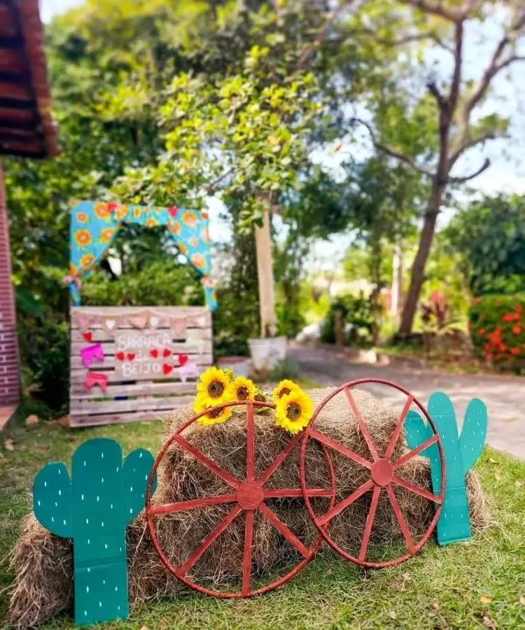 Decoração rústica de festa Junina ao ar livre com fardos de feno, rodas de carroça, cactos de madeira e girassóis. Ao fundo, uma barraquinha decorada com a frase "Barraca do Beijo".