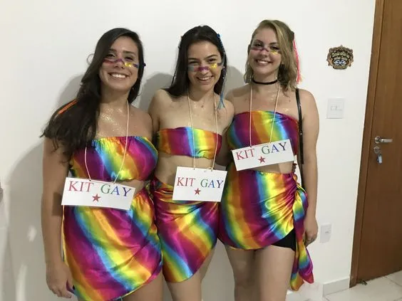 Três mulheres de pele clara com top e saia coloridos e placa "kit gay"