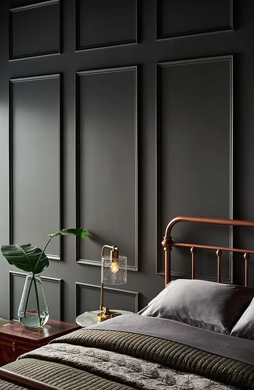 Quarto com roupa de cama cinza, cabeceira de cobre, cômoda de madeira, parede escura com moldura 
