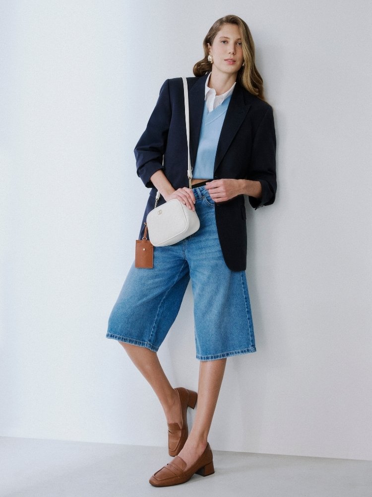 Modelo usando camisa azul, blazer marinho, bermuda jeans, bolsa branca e mocassim marrom da linha Office da Anacapri